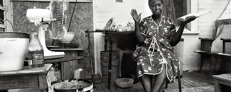 Black and white photograph of an elderly African American woman sitting on a cluttered porch, holding up a Bible in her left hand and holding up her right hand as if testifying.